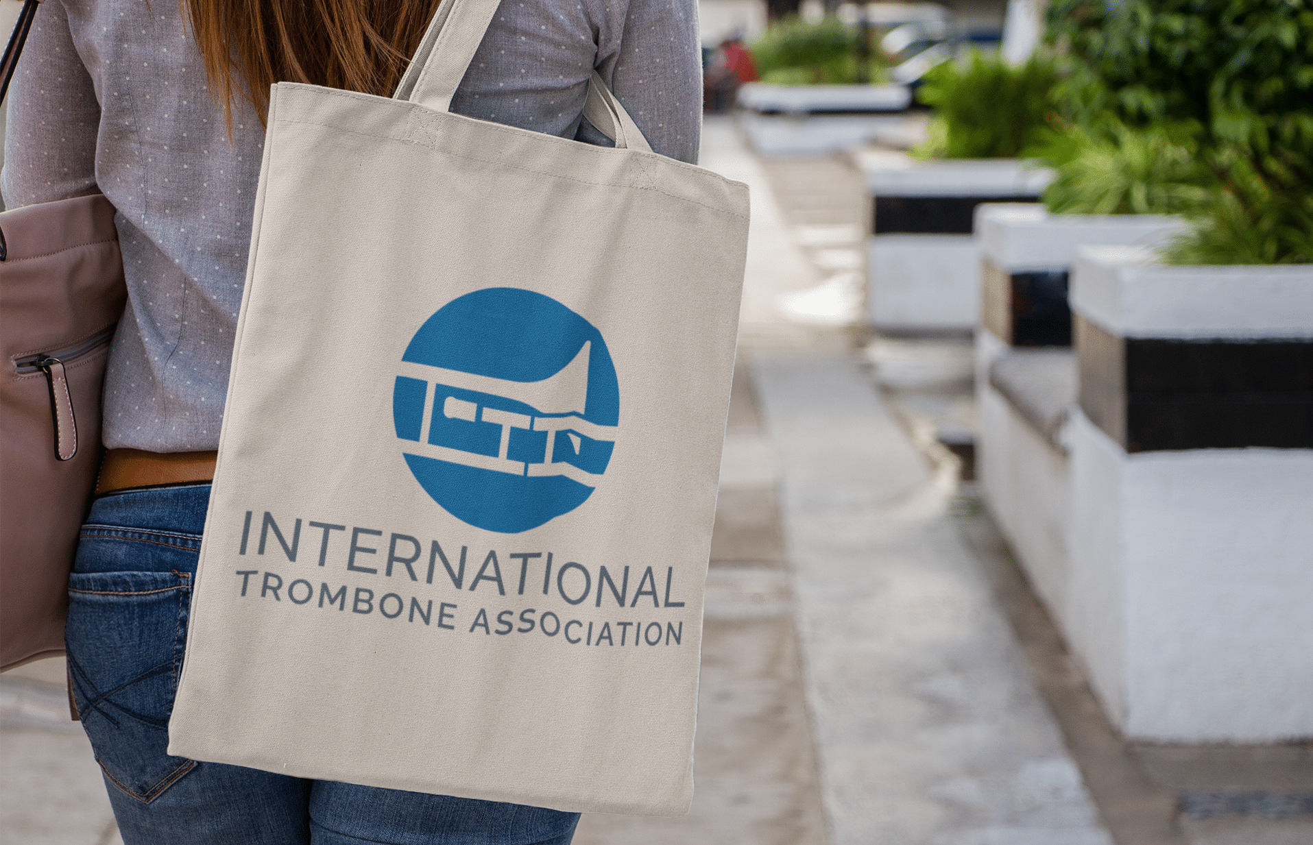 A person walks outdoors in Richmond VA carrying a beige tote bag with the blue “International Trombone Association” logo, a stylized trombone inside a circle—a great example of eye-catching logo design.