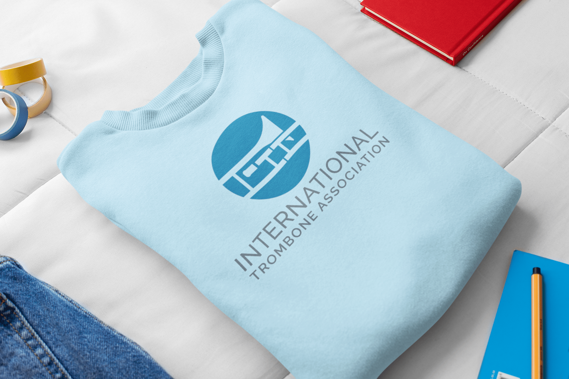 A light blue sweatshirt with the International Trombone Association logo design, featuring a stylized trombone in a blue circle, is neatly folded on a white surface near a red notebook, pen, and yellow tape.