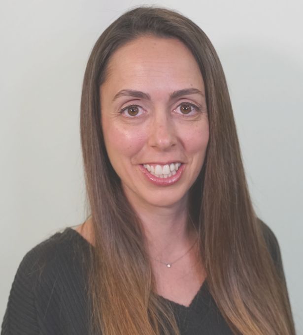 A woman with long straight brown hair and brown eyes smiles at the camera. She is wearing a dark sweater and a delicate necklace, standing in front of a plain light background, representing Moonlit Media, a creative agency.
