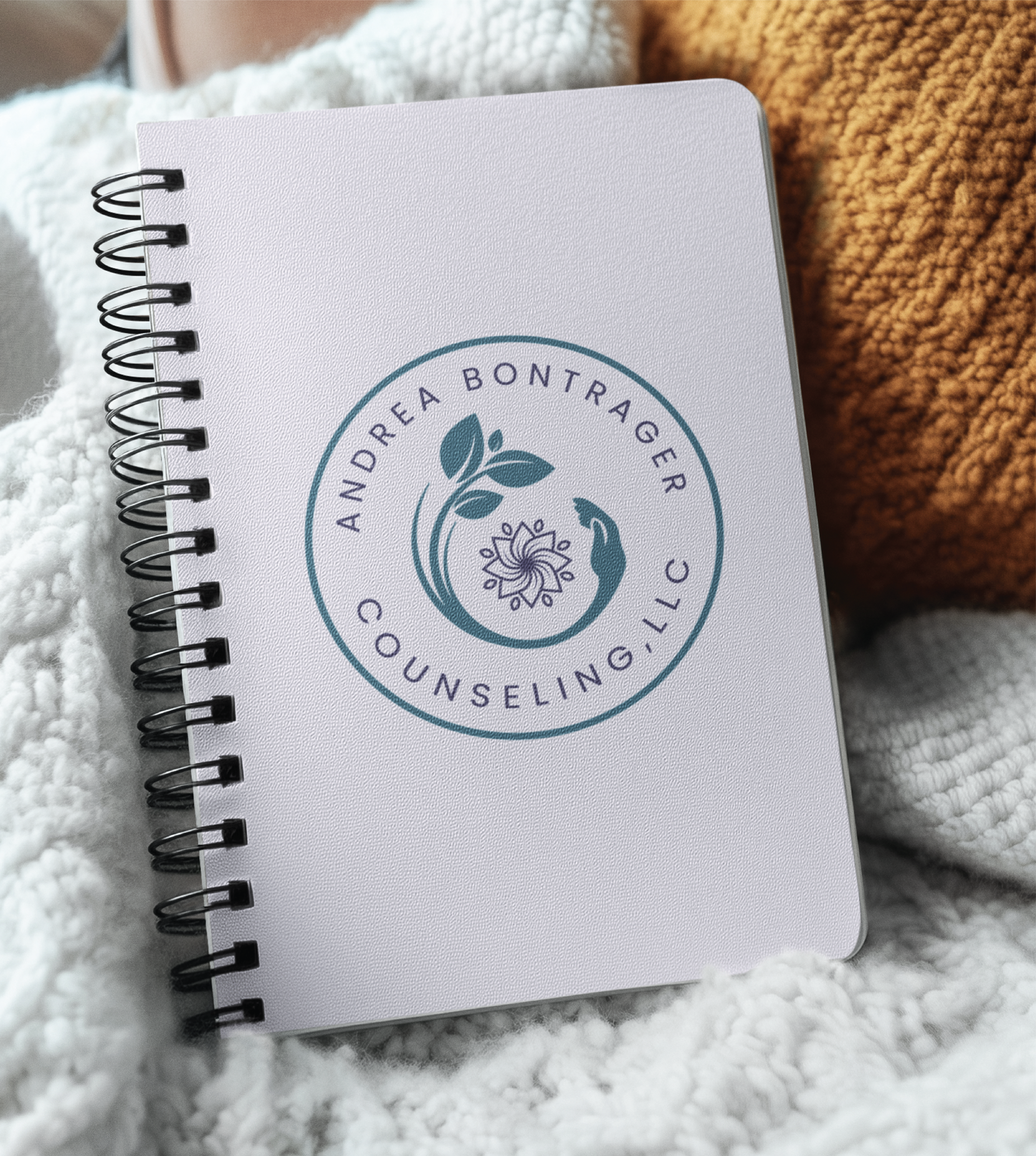 A spiral-bound notebook with a custom logo design reading “Andrea Bontrager Counseling, LLC,” featuring a leafy branch and two hands forming a circle, rests on a cozy white knit blanket beside a textured orange pillow.