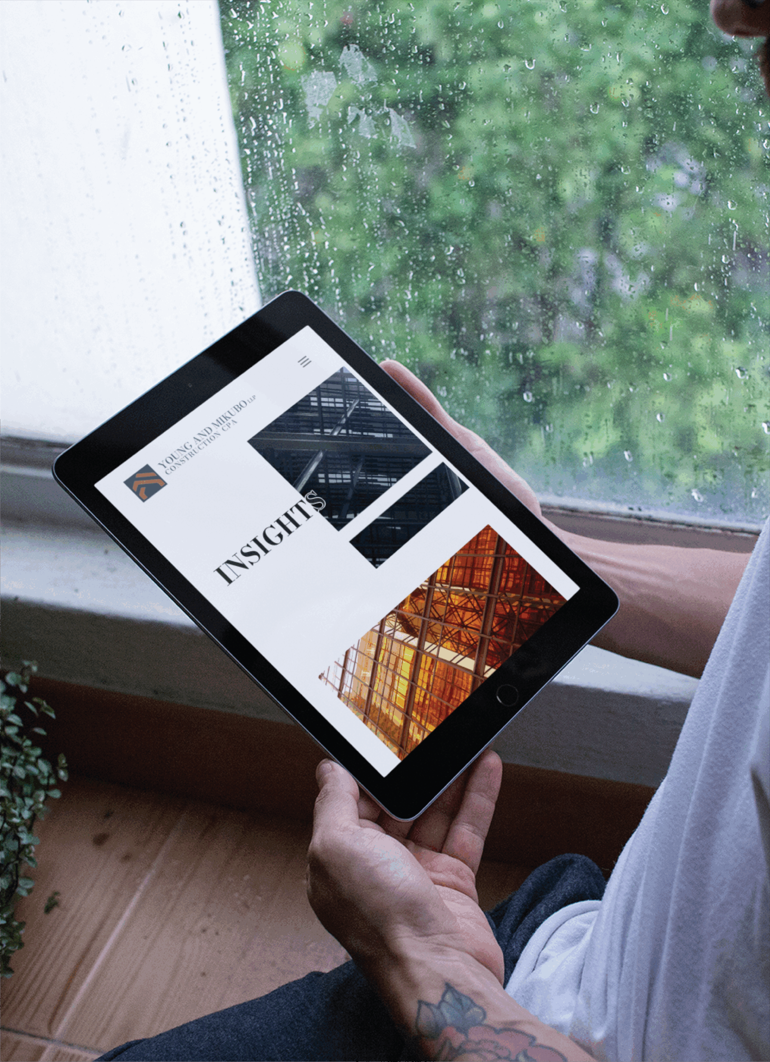 A person holding a tablet displaying a roofing company’s website design with architectural images and the word INSIGHTS, sitting by a rainy window.