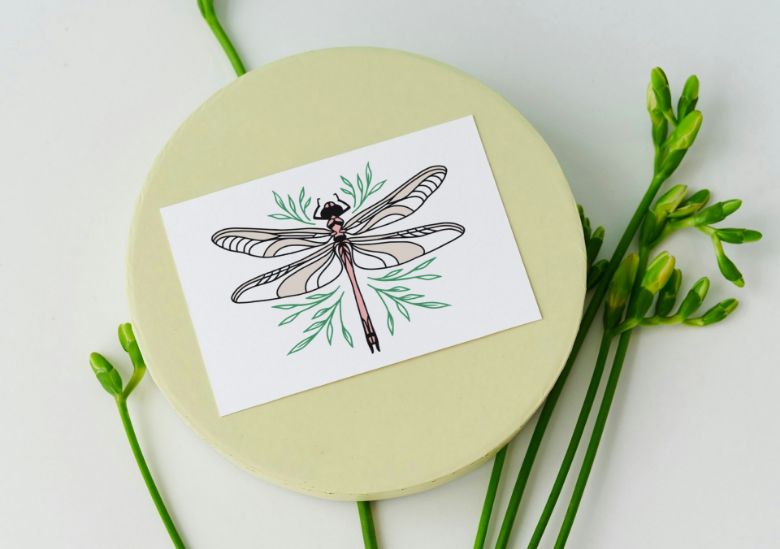 A card featuring a delicate dragonfly illustration with green leaves is placed on a round, pale yellow surface, making it a perfect inspiration for small businesses seeking custom logo design, with green flower stems arranged beside it.