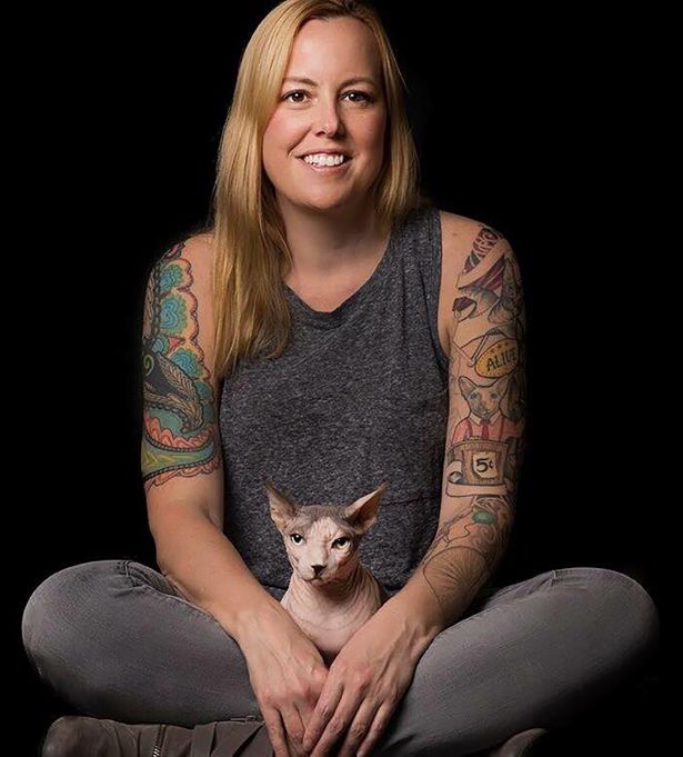 A smiling, woman-owned web design entrepreneur with long blonde hair and colorful arm tattoos sits cross-legged in a sleeveless gray top and jeans, holding a hairless Sphynx cat in her lap against a black background.