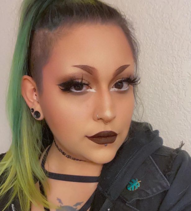 A woman-owned branding expert with long green hair, dramatic makeup, dark lipstick, and piercings poses in a black jacket and choker before a light textured wall.