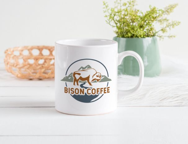A white mug with a Bison Coffee logo featuring a bison and mountains sits on a white surface. Showcasing thoughtful logo design, the scene includes a woven basket and a green vase with small leafy plants in the background.