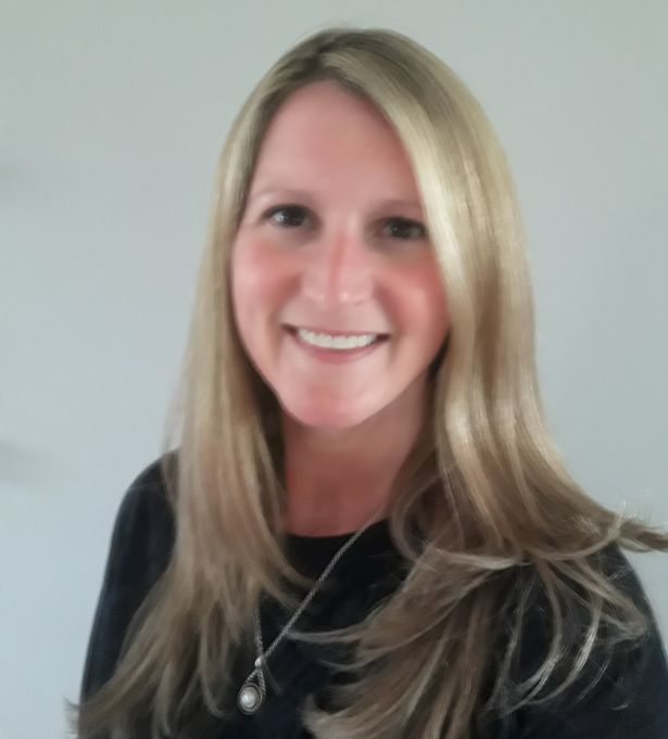 A woman with long, straight blonde hair and a black top smiles at the camera. She is wearing a necklace and stands against a plain light background, representing her woman-owned web design and branding business.