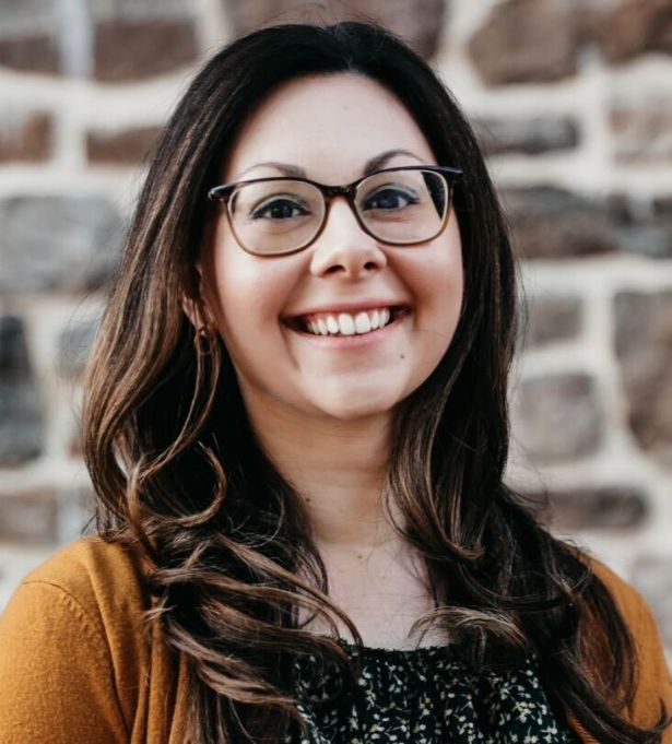 A woman with long brown hair, wearing glasses and a mustard-colored sweater, smiles in front of a stone wall background—proudly representing her woman-owned branding studio.