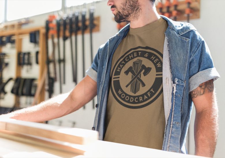A man in a denim shirt and tan Hatchet & Hew Woodcraft t-shirt stands in a bright woodworking shop—an inspiring space for entrepreneurs and small businesses, with tools and wooden boards in the background.
