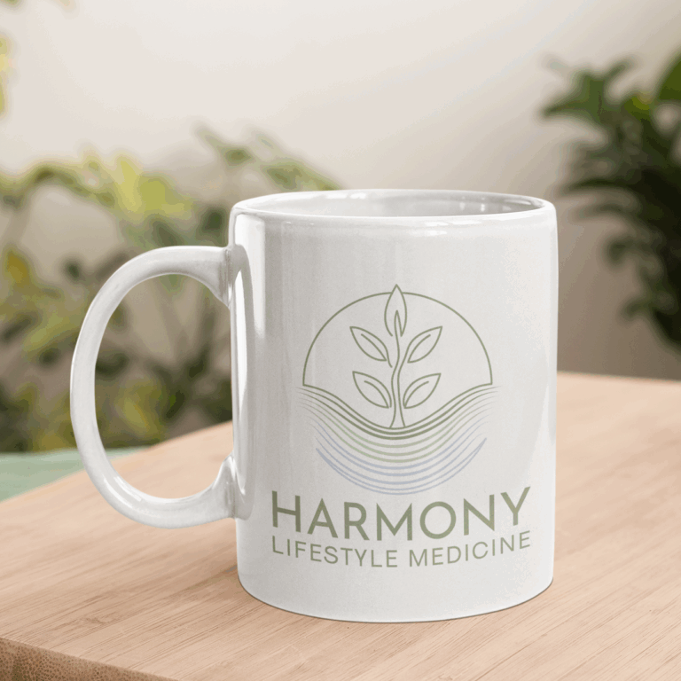 A white mug on a wooden surface, featuring a green leaf logo above wavy lines and the text HARMONY LIFESTYLE MEDICINE, highlights wellness branding. Blurred green plants are visible in the background.