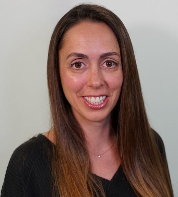A woman with long brown hair and brown eyes is smiling at the camera. Wearing a black top and delicate necklace, she poses against a light background, capturing the essence of woman-owned branding.