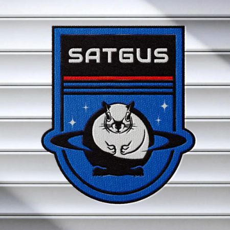 A blue SATGUS patch, perfect for branding, shows a black-and-white rodent inside a ringed planet with stars behind and bold red, white, and black stripes above. The patch rests on a white, ridged surface.