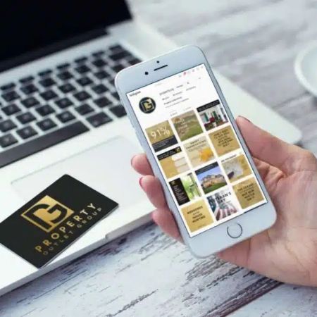 A person holds a smartphone displaying a real estate-themed social media page, next to a laptop and a black and gold Property Power Group business card—showcasing modern branding and website design on a light wooden desk.