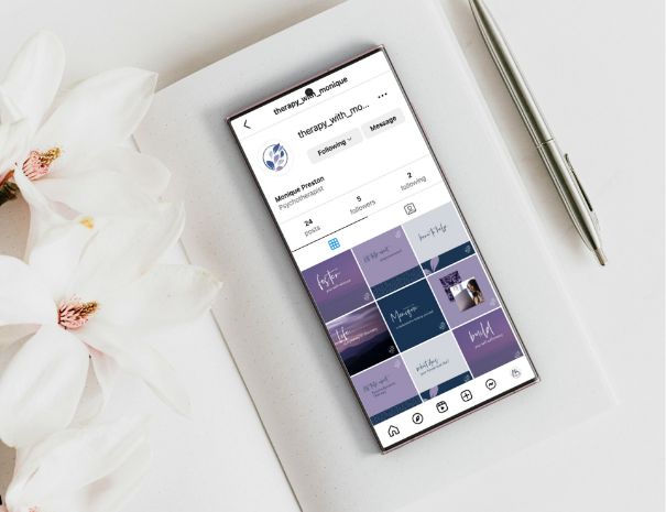 A smartphone displaying an Instagram profile lies on a white notepad beside a silver pen and white flowers, creating a clean, elegant workspace scene ideal for website design or sharing your brand story.