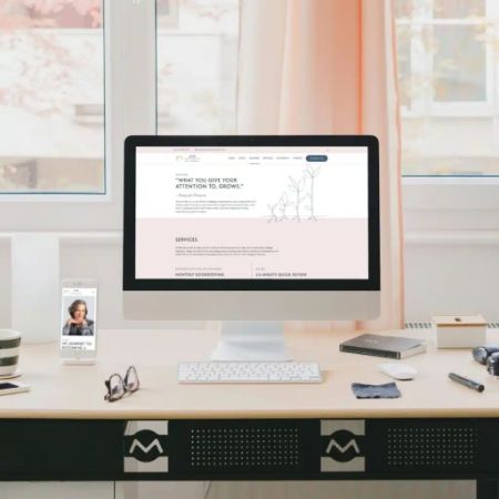 A minimalistic desk setup with a computer displaying a website design, a smartphone on a stand, a notepad, pen, and glasses. The desk sits by a window with light curtains and autumn trees visible outside—perfect for creative support.