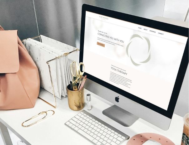 A neatly organized white desk with a computer displaying a yoga therapy website design, a peach bag, gold pen holder with pens, keyboard, mouse, marble file holders with folders, and paper clips.