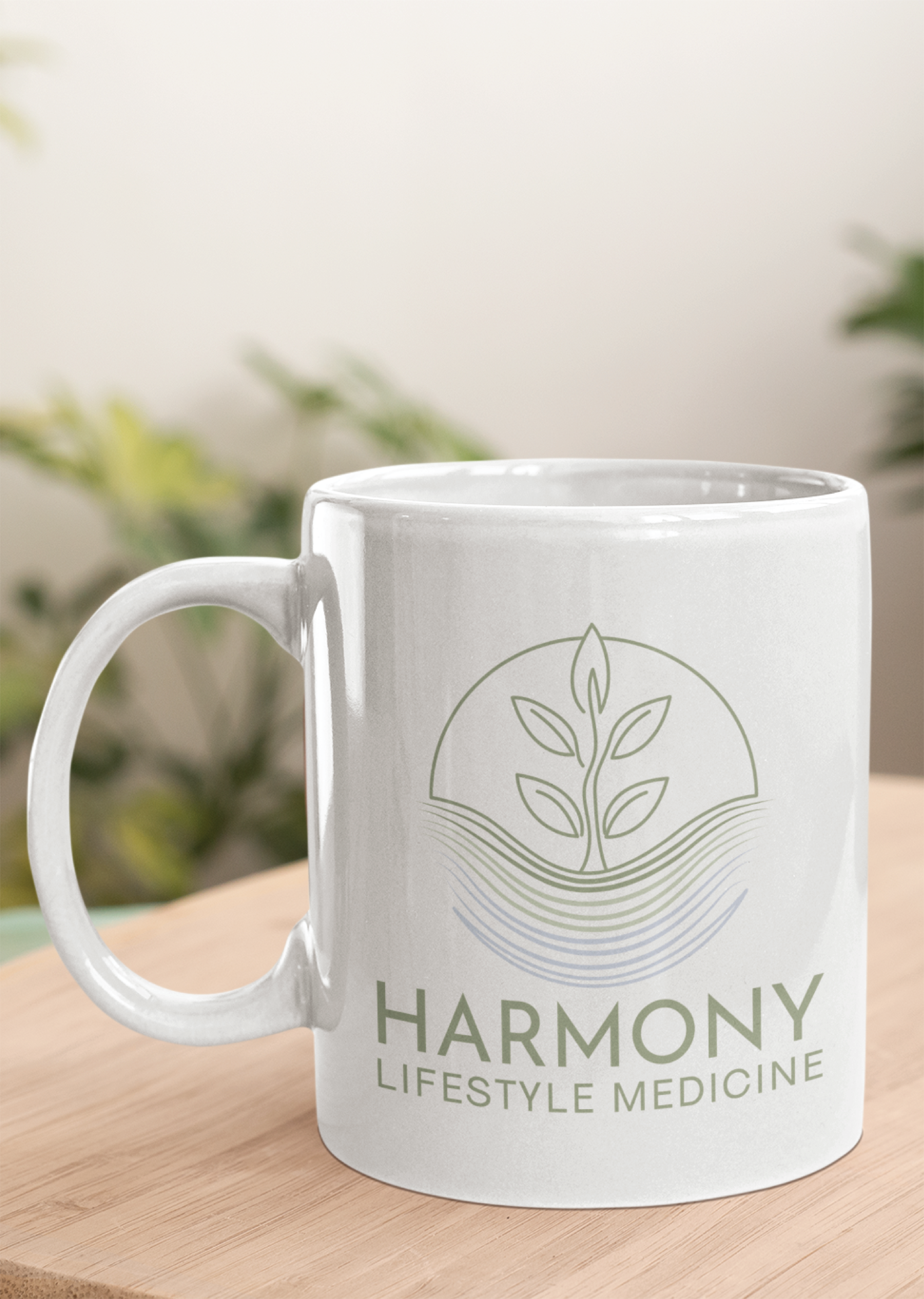 A white mug on a wooden surface features a green plant logo above blue waves and the words Harmony Lifestyle Medicine, showcasing thoughtful branding. Blurred green plants are visible in the background.