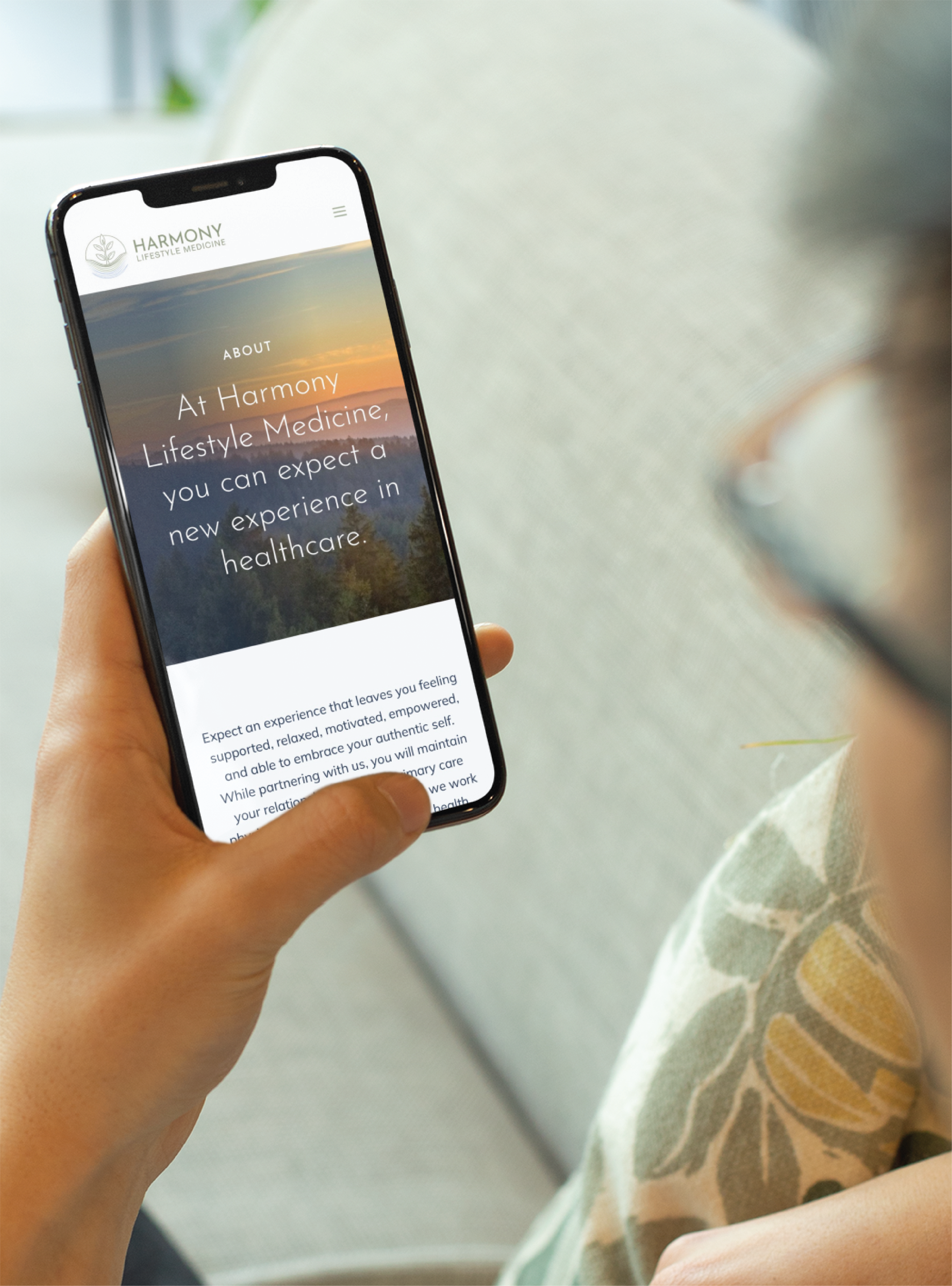 A person holding a smartphone, reading a website about Harmony Lifestyle Medicine. The screen displays text on branding and healthcare innovation, with a serene sunset landscape background—showcasing thoughtful website design.