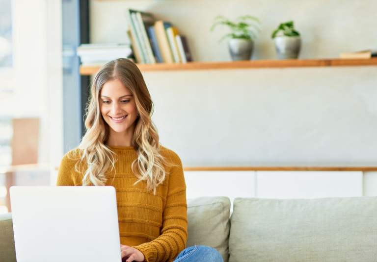 A woman with long blonde hair, wearing a mustard yellow sweater, sits on a light-colored sofa and smiles while using her laptop, likely working as a web designer. Books and potted plants decorate the shelf in the background.
