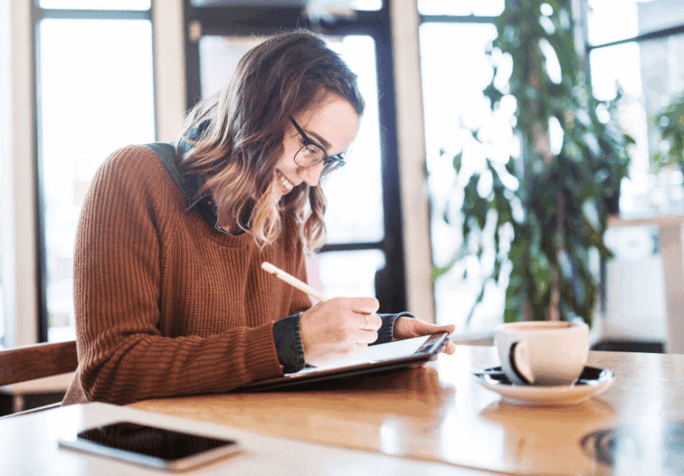 A smiling woman wearing glasses and a brown sweater writes on a digital tablet at a café table, brainstorming ideas for custom logo design. A coffee cup, smartphone, and green plants add charm to the cozy, naturally lit setting.