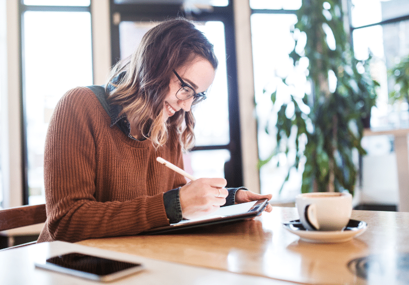 A smiling woman wearing glasses and a brown sweater writes on a digital tablet at a café table, brainstorming ideas for custom logo design. A coffee cup, smartphone, and green plants add charm to the cozy, naturally lit setting.