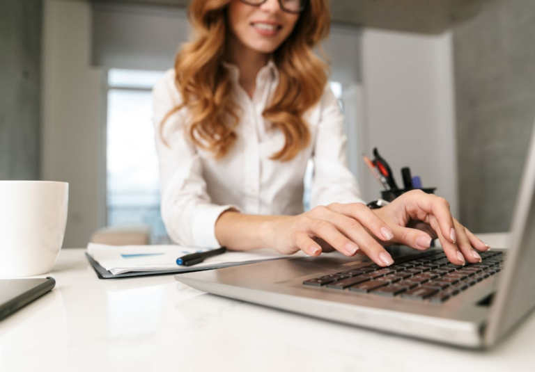A woman with long red hair and glasses types on a laptop at a desk with a notebook, pen, and coffee cup. She is smiling, working on logo design services for Moonlit Media in a modern office setting.