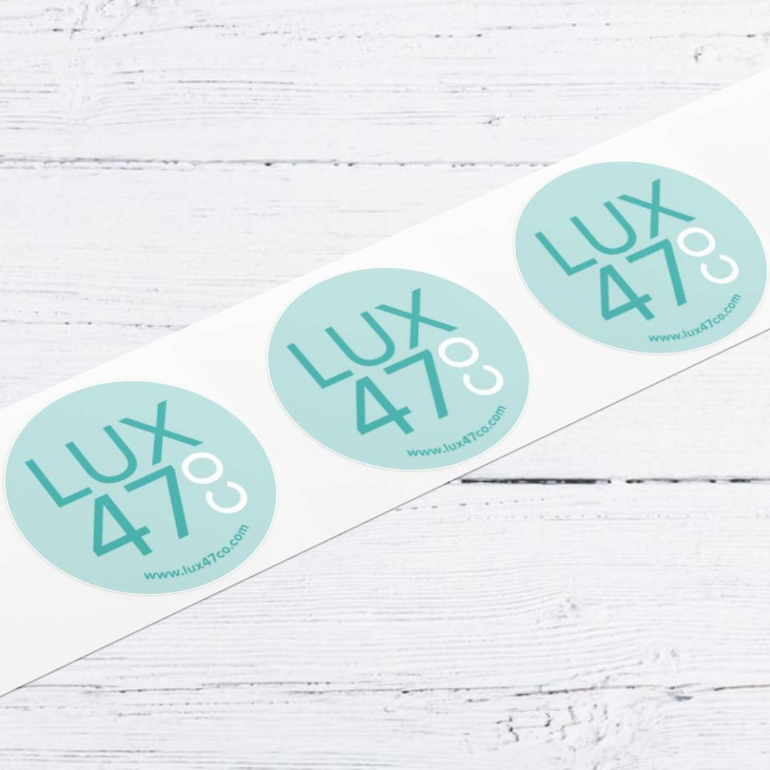| Moonlit Media LLC A white strip with three round, light blue stickers that read LUX 47° in bold text, placed on a white wooden surface. Highlight your brand with moonlit media’s logo design services. The website www.lux47co.com appears on each sticker.
