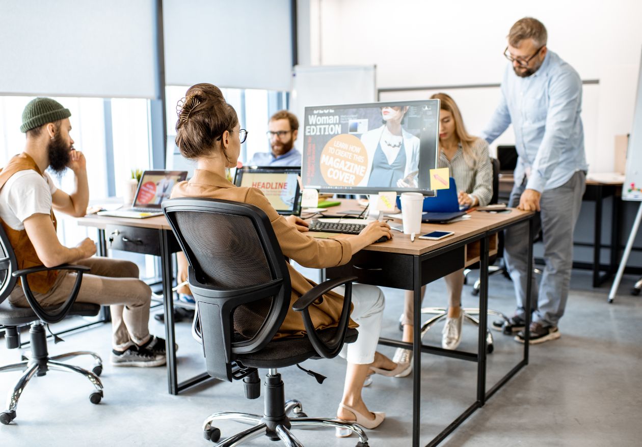 A group of six people work together in a modern office, sitting at desks with computers and magazines. A web designer stands, presenting custom logo design ideas to the others, who appear focused and engaged in discussion.
