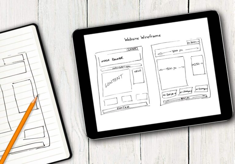 A tablet displaying a hand-drawn website wireframe sits on a white wooden surface next to an open notebook with sketches and a pencil, reflecting the creative process behind moonlit media's custom website design solutions.