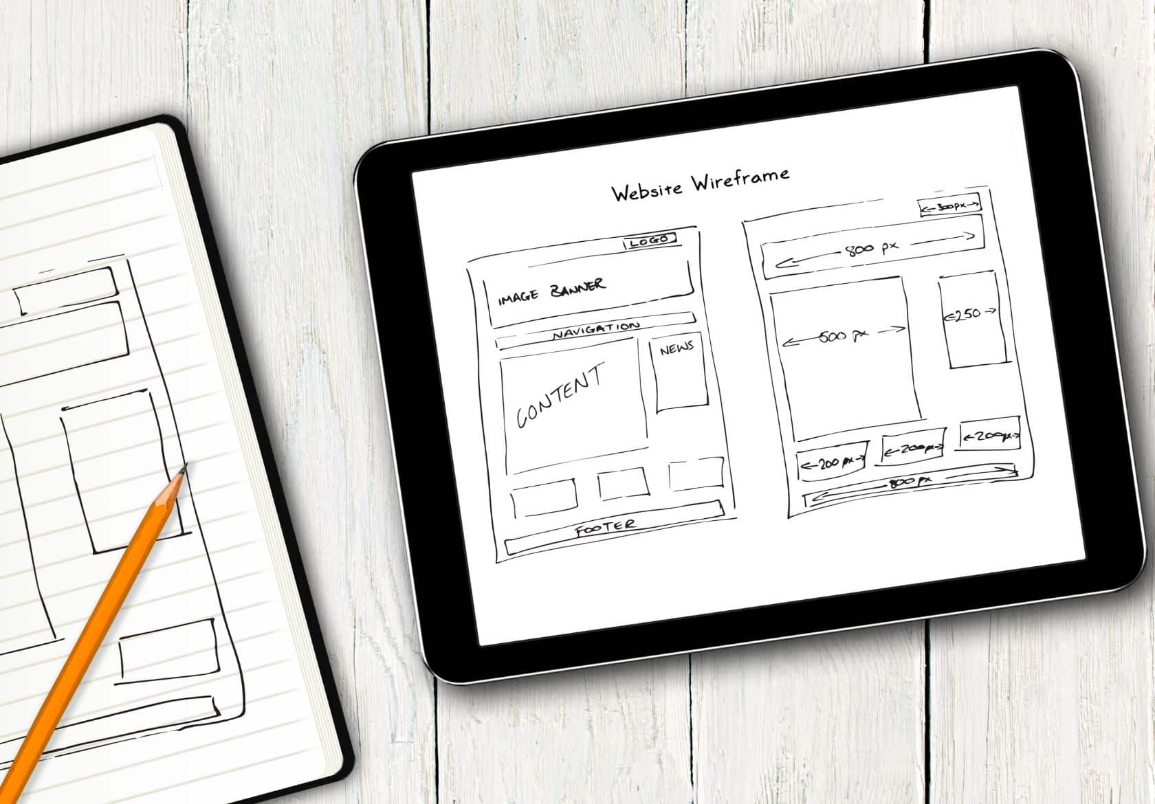 A tablet displaying a hand-drawn website wireframe sits on a white wooden surface next to an open notebook with sketches and a pencil, reflecting the creative process behind moonlit media's custom website design solutions.
