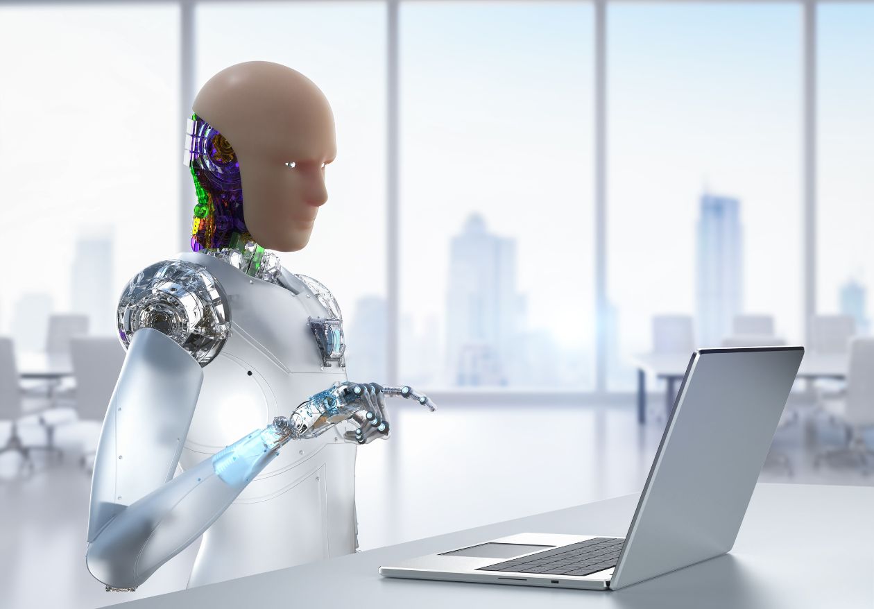 A humanoid robot with exposed circuitry is sitting at a desk in a modern office, using a laptop—perhaps working on custom website design. Large windows reveal a blurry cityscape in the background.