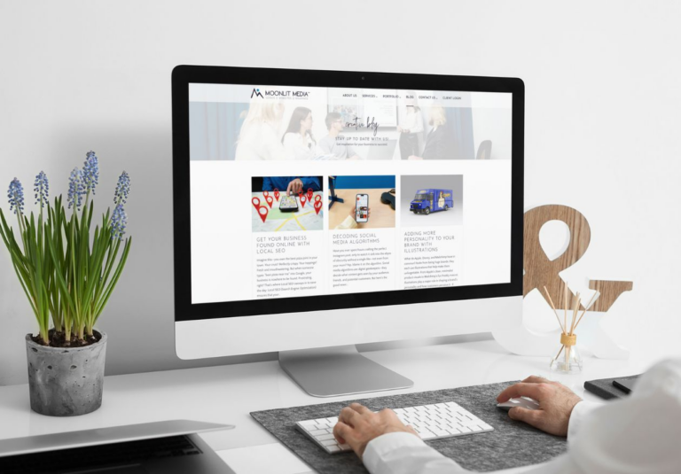 A web designer uses a desktop computer at a modern white desk with blue potted flowers, a keyboard, mouse, and office decor. The computer screen displays a business website featuring custom website design with images and text.