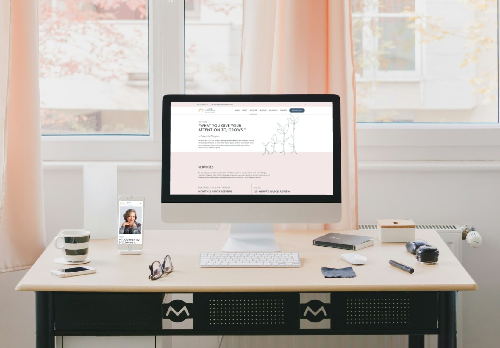A modern desk with a computer, smartphone, keyboard, mouse, coffee mug, and notepad. The computer screen displays a custom website design, while the window behind the desk shows trees with autumn leaves.