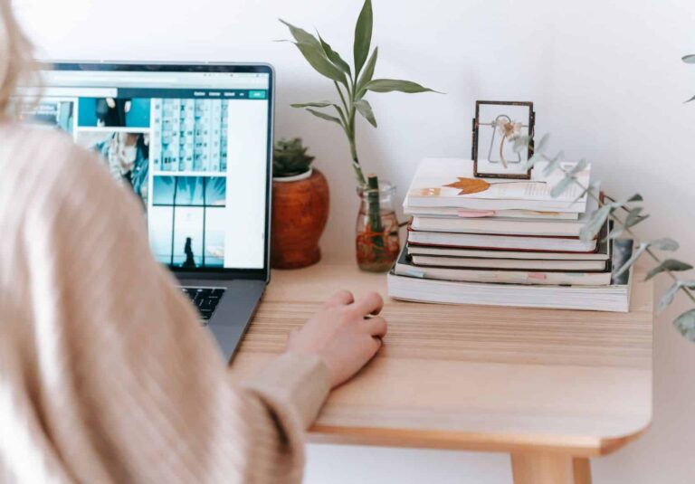 A web designer uses a laptop at a wooden desk with stacks of books, a small plant, and a framed picture. The workspace is bright and neatly organized, perfect for brainstorming custom logo design ideas.