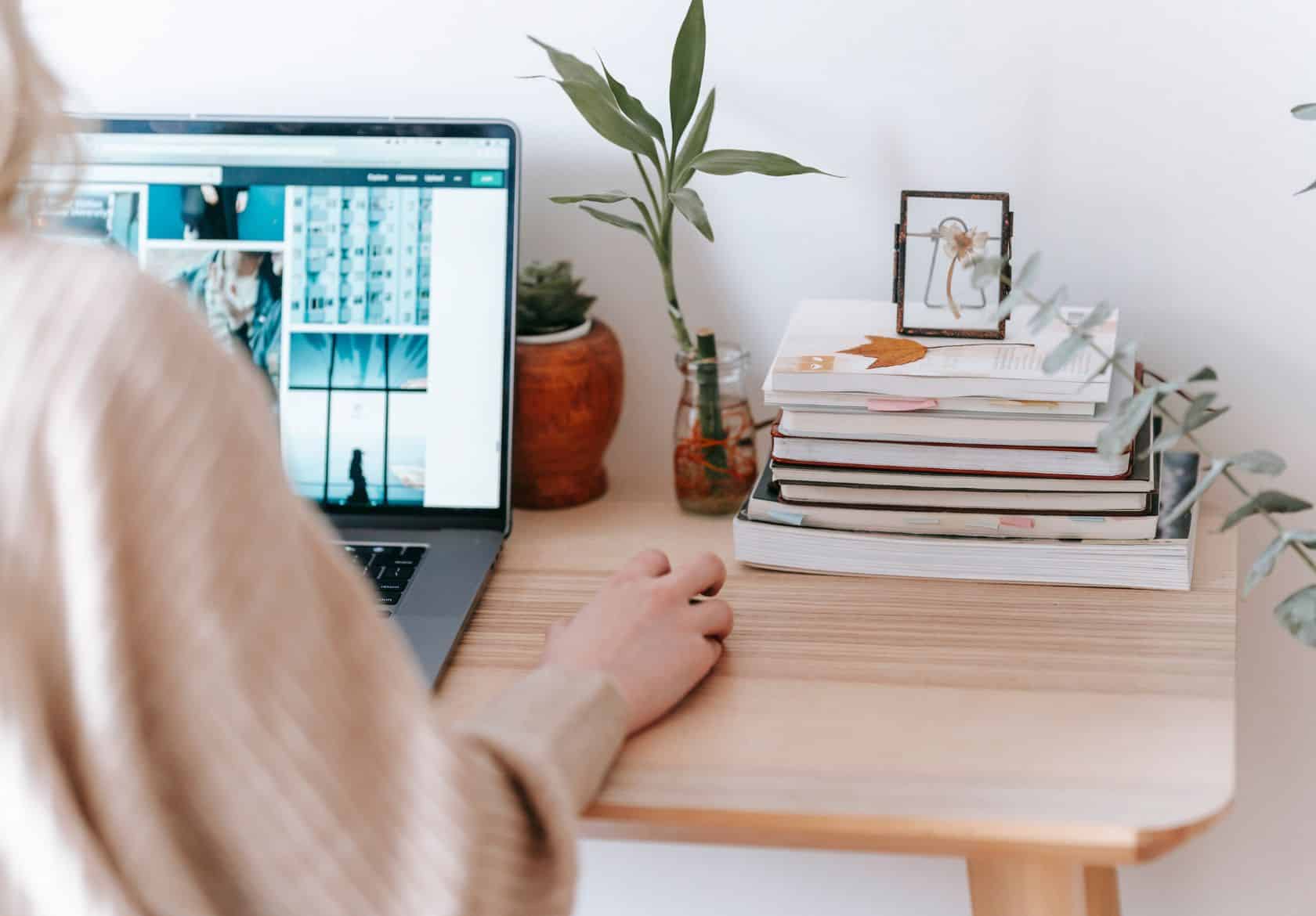 A web designer uses a laptop at a wooden desk with stacks of books, a small plant, and a framed picture. The workspace is bright and neatly organized, perfect for brainstorming custom logo design ideas.
