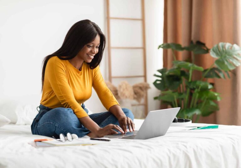 A woman sits cross-legged on a bed, smiling while using her laptop to browse web design services. She wears a yellow top and jeans, with books, a phone, and a pen nearby. The background features plants and neutral decor.