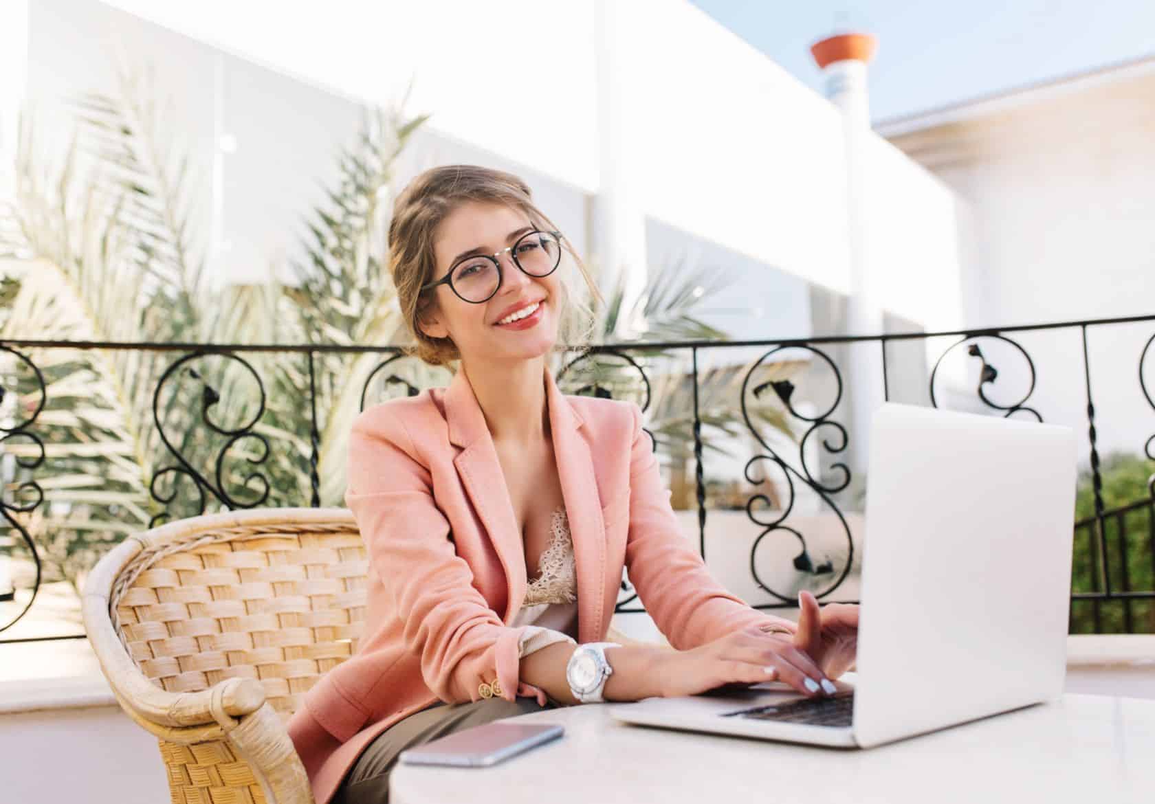 A smiling woman with glasses in a pink blazer sits outdoors at a wicker table, typing on her laptop. She offers custom website design while her smartphone rests nearby, with leafy plants and a building in the background.