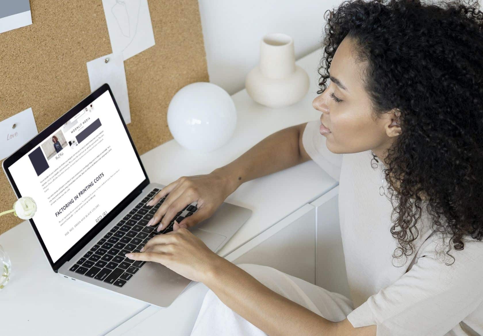 A woman with curly hair sits at a white desk using a laptop, appearing focused on custom website design. The desk holds a vase, lamp, and papers with a corkboard on the wall in the background.