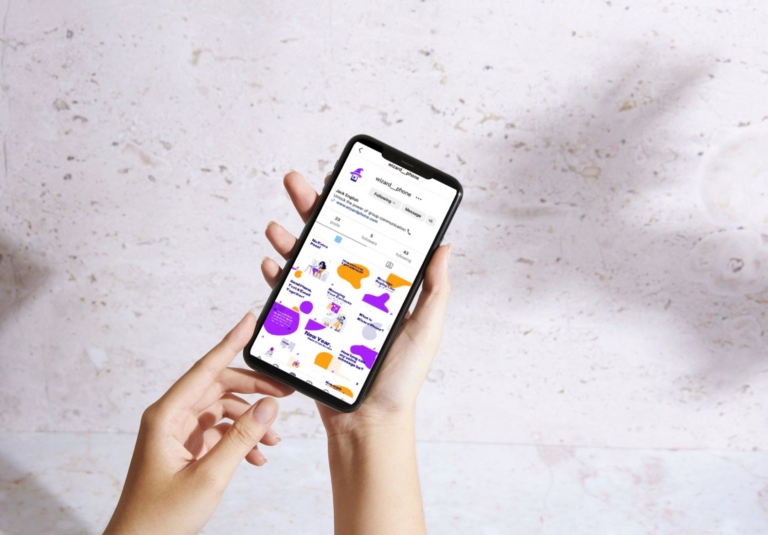 A person holds a smartphone displaying a social media profile with posts featuring colorful speech bubbles and illustrations, showcasing custom logo design. The phone is held with both hands over a pale, textured surface.