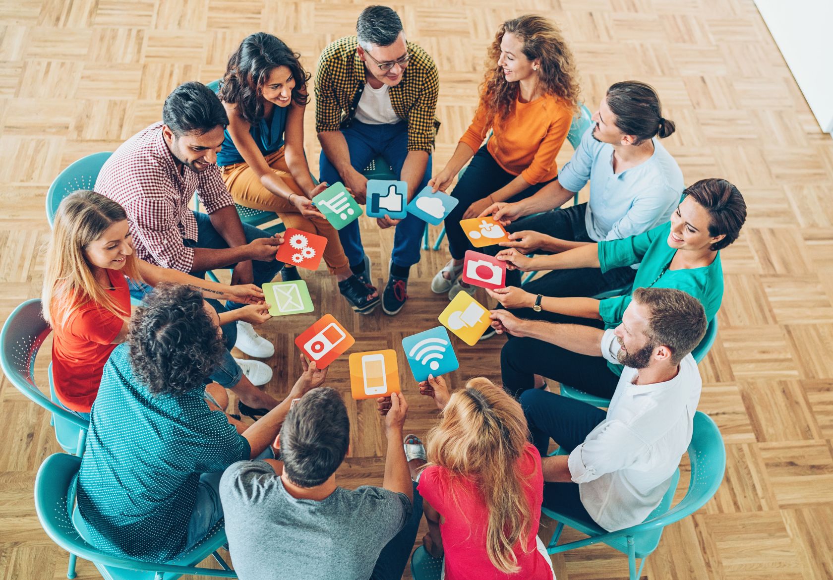 A group of people sitting in a circle, each holding colorful icons representing technology, communication apps, and custom website design, in a bright room with wooden flooring.