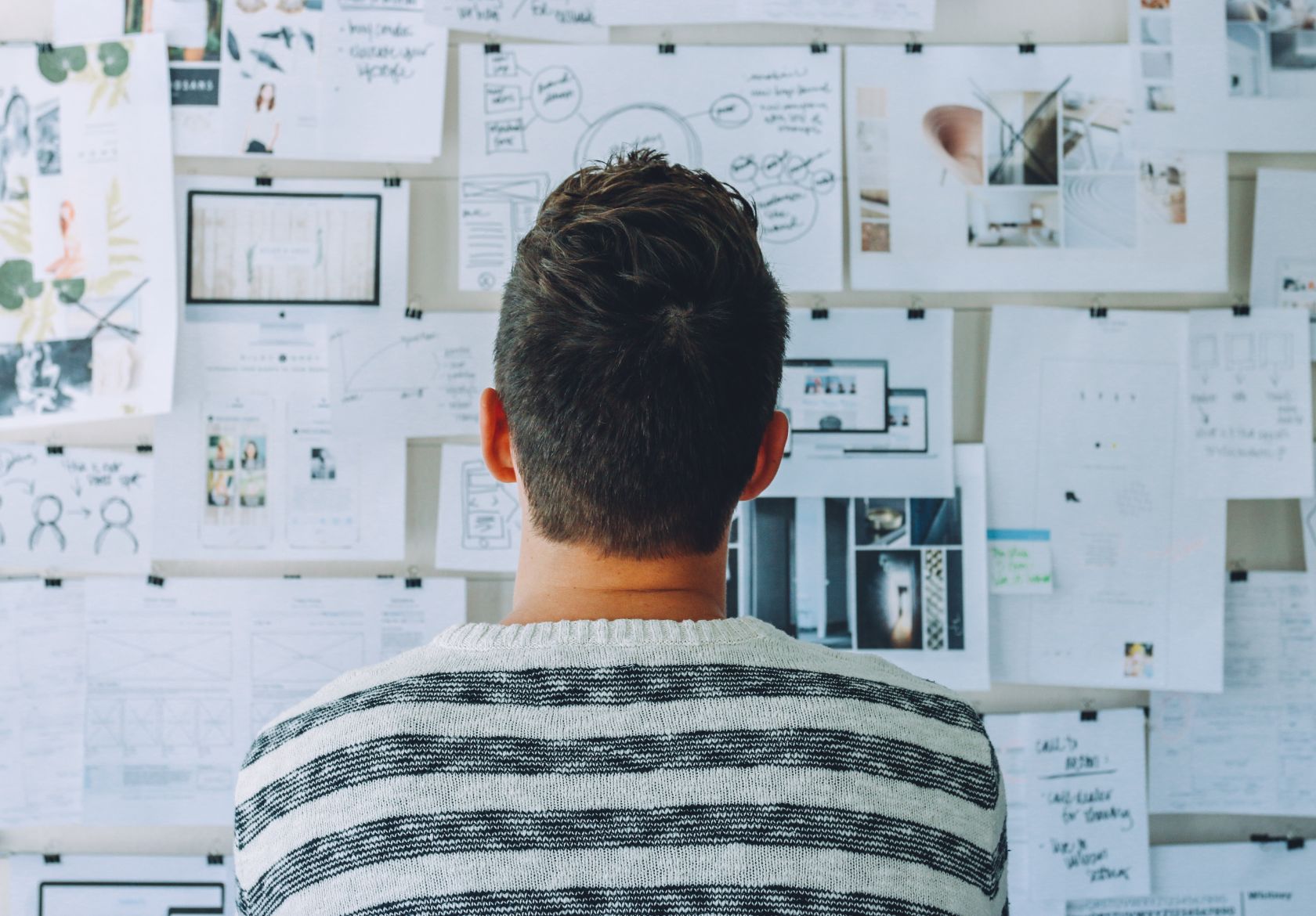 A person with short brown hair, wearing a striped sweater, faces a wall covered with papers and diagrams—perhaps brainstorming ideas for custom website design or logo design services at Moonlit Media.