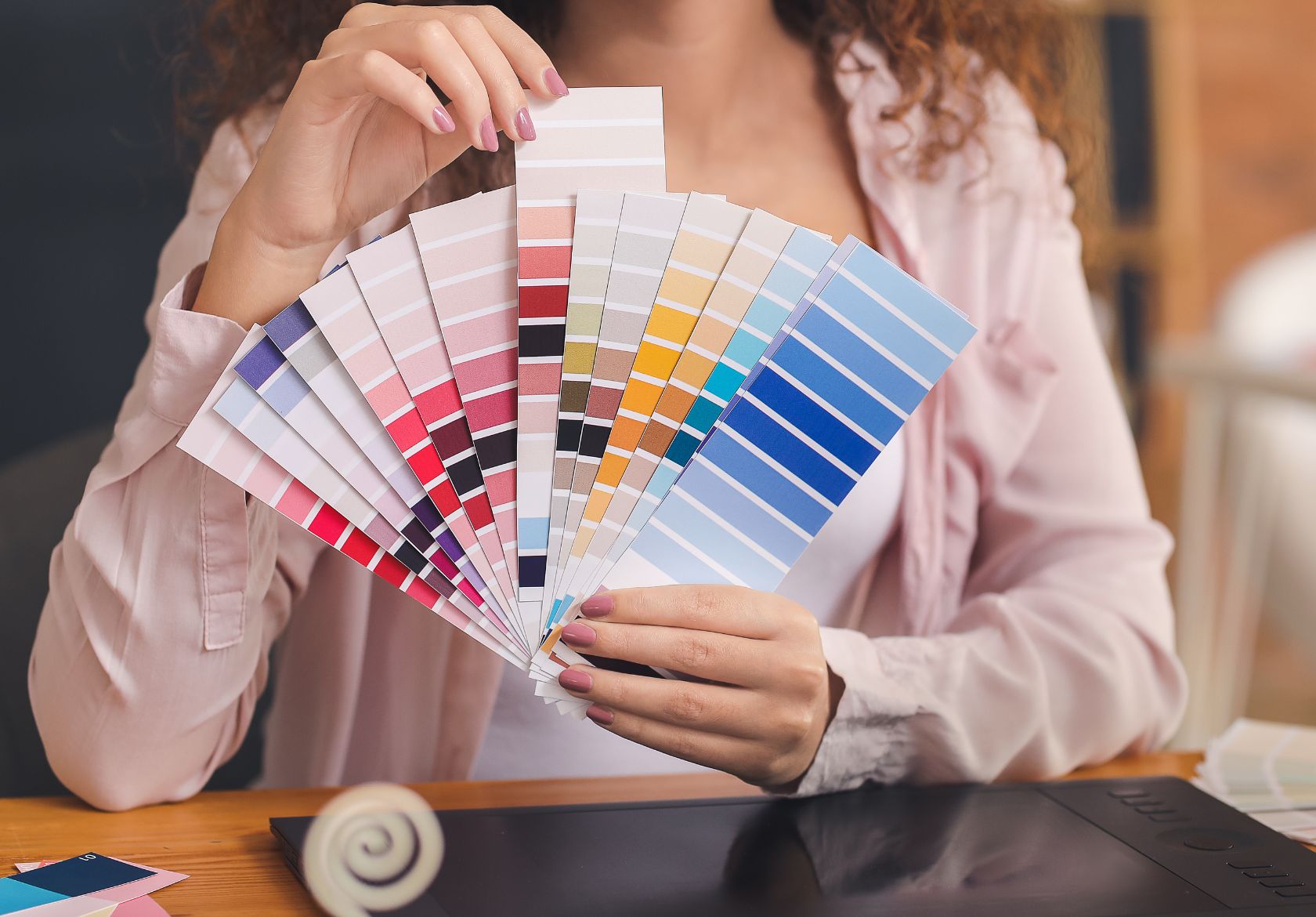 A person holding a fan of color swatches, displaying a wide variety of shades from red and pink to yellow, green, and blue. Seated at a desk in a light pink shirt, they explore vibrant palettes for Moonlit Media’s web design services.