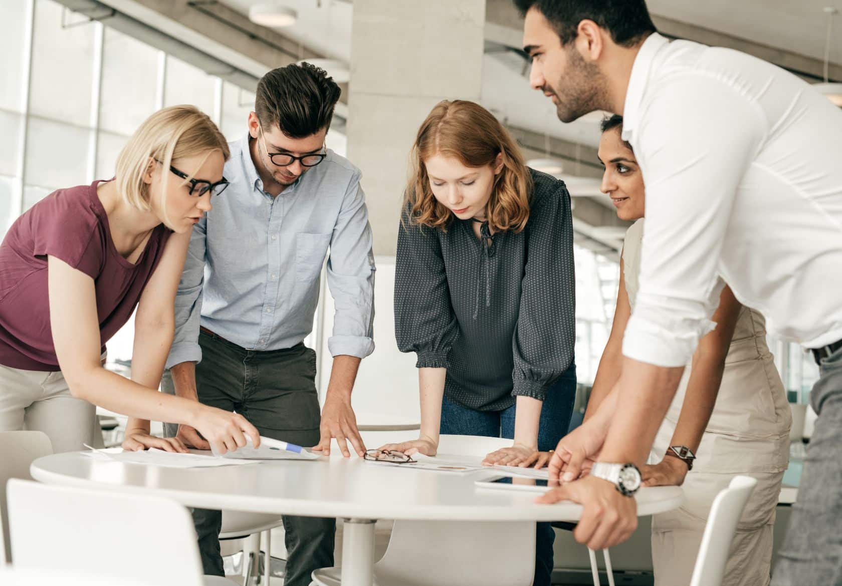 Five people stand around a table in a modern office, closely collaborating on custom website design and looking at documents from Moonlit Media, appearing focused and engaged in discussion.