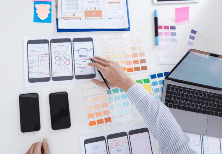 A person designs a mobile app user interface using printed wireframes, color swatches, and a laptop. Custom logo design samples and two smartphones are spread out on the white desk alongside notes and design tools.