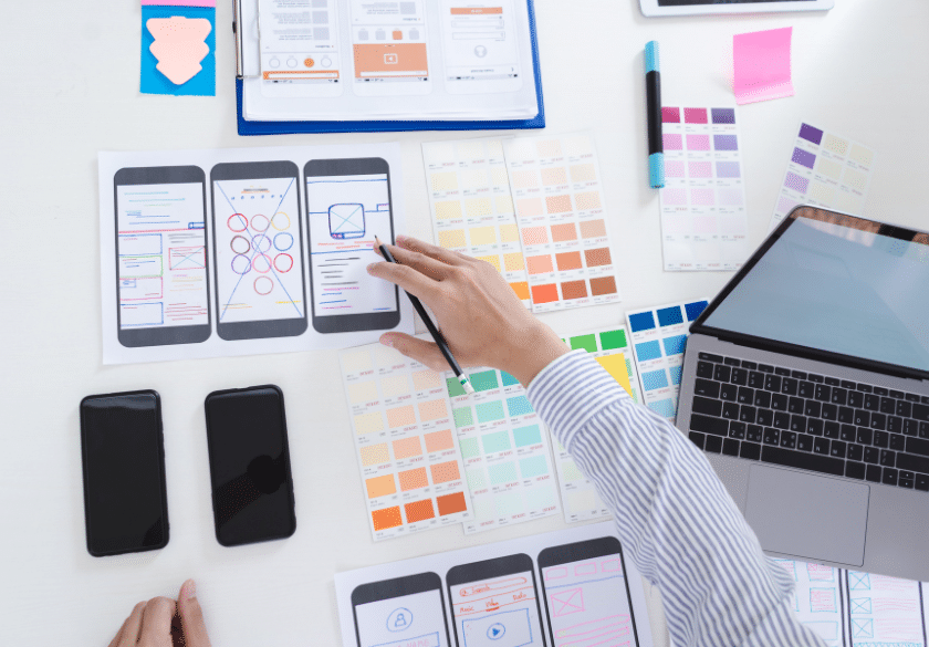 A person designs a mobile app user interface using printed wireframes, color swatches, and a laptop. Custom logo design samples and two smartphones are spread out on the white desk alongside notes and design tools.