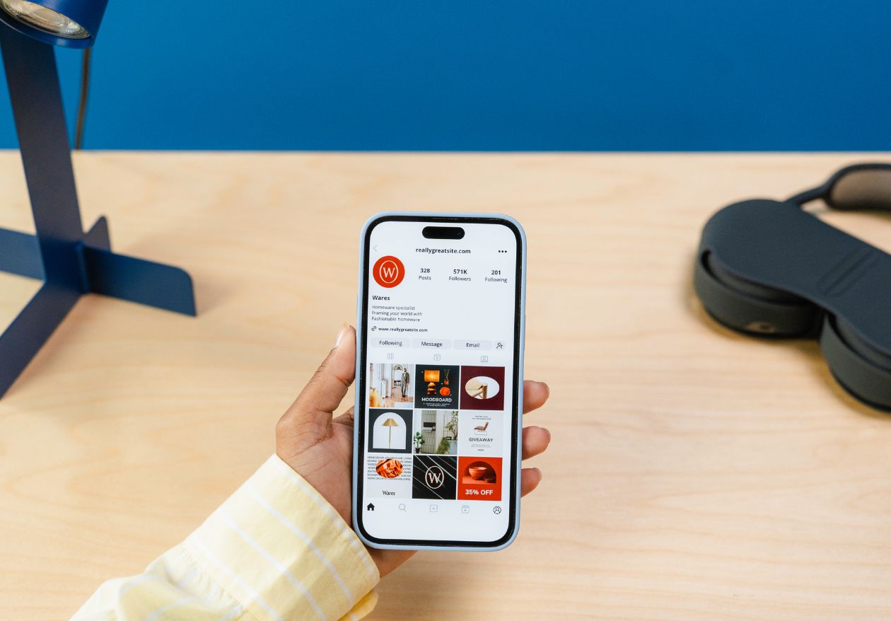A person holding a smartphone displaying an Instagram profile with design-related posts; the background shows a wooden desk, part of a lamp, and a pair of black headphones—perfect for finding moonlit media’s custom website design inspiration.