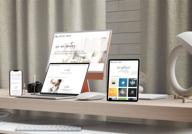 A modern workspace with a smartphone, laptop, tablet, desktop computer, wireless mouse, and lamp on a wooden desk—all showcasing Moonlit Media’s web designer expertise and branding—set against beige curtains.