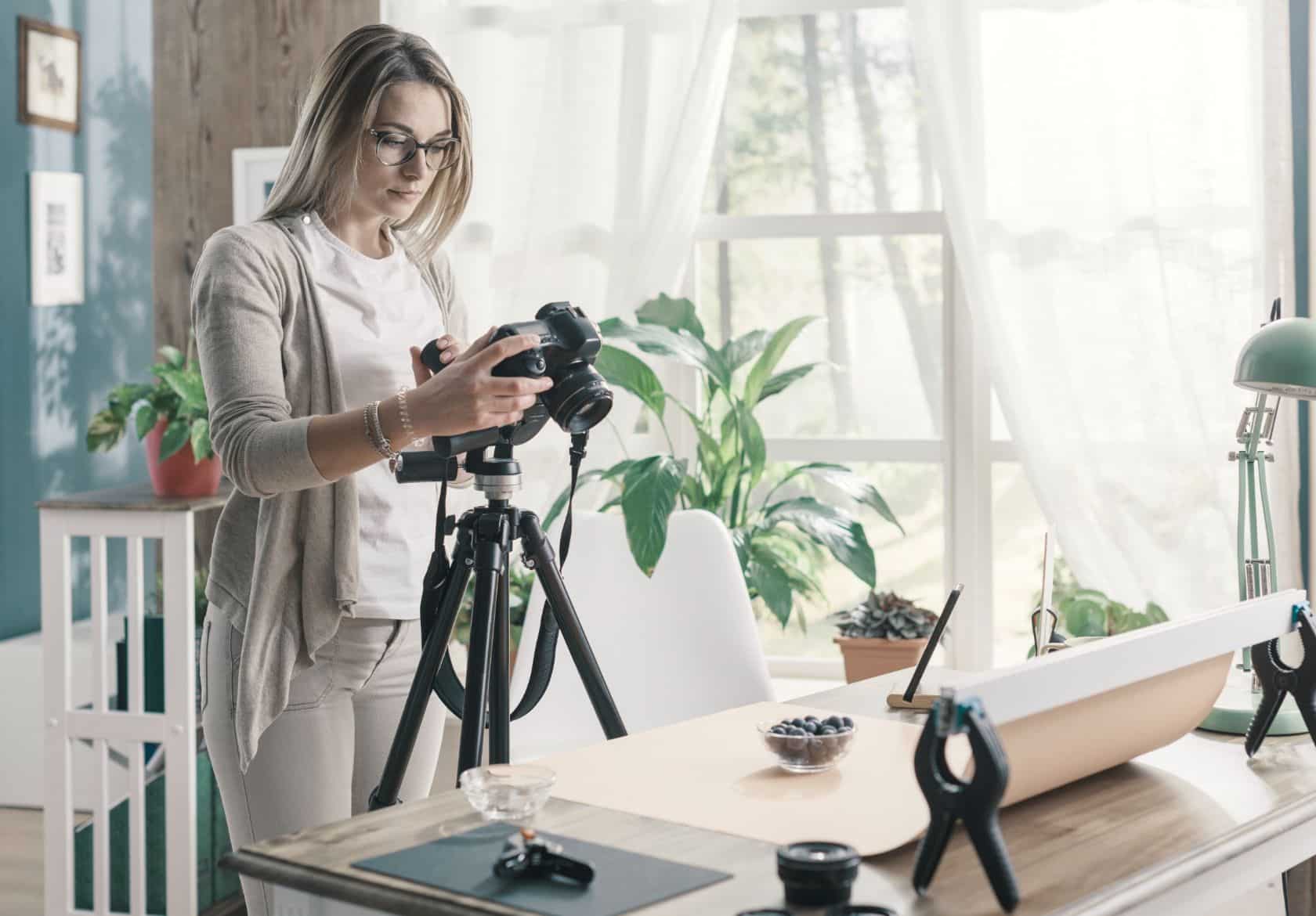 A woman adjusts a camera on a tripod in a bright, plant-filled room, preparing to photograph a plate of food—perfect inspiration for any web designer seeking fresh visuals for custom website design projects.