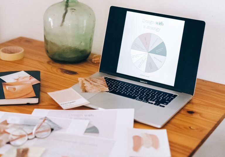 A laptop on a wooden desk displays a pie chart titled Design with a strategy. Nearby are papers, a notebook, glasses, a fabric swatch, and a large green glass vase—capturing the creative workspace of a web designer at Moonlit Media.