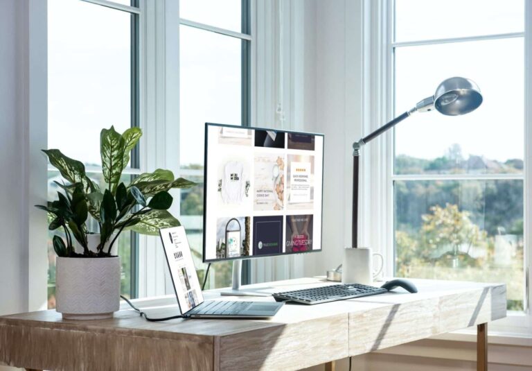 A bright home office with a large window, a wooden desk set up for custom website design work, a computer monitor, laptop, keyboard, potted plant, desk lamp, and white mug. Greenery is visible outside the window.