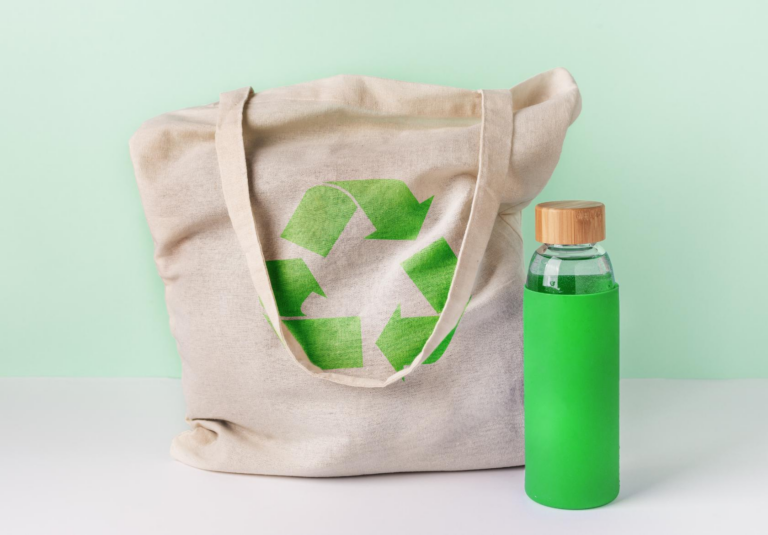 A beige tote bag with a green recycling symbol sits next to a reusable glass water bottle, both placed on a white surface—showcasing Moonlit Media’s stylish flair, perfect for those seeking custom website design or logo design services.